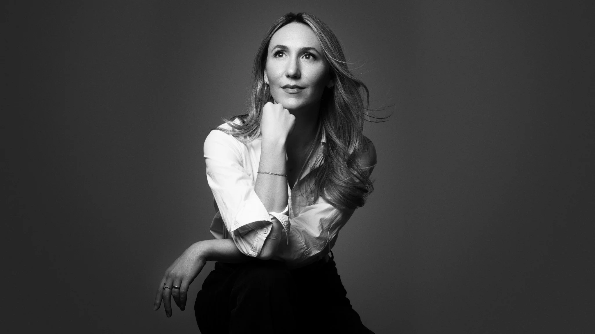 A woman with long wavy hair rests her chin on her hand in a black-and-white studio portrait.