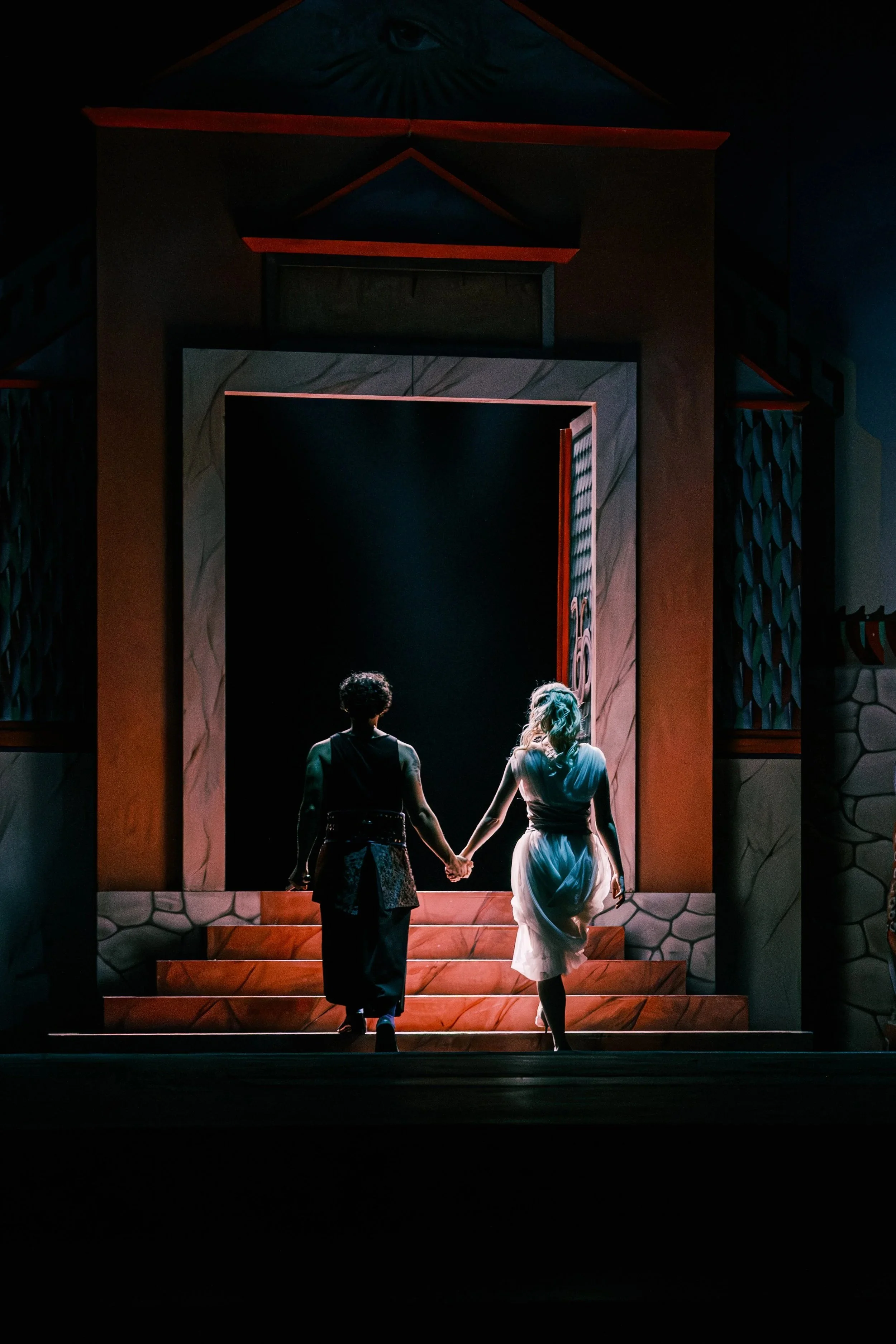 Two actors holding hands, walking up stairs on a stage designed like a theatrical set, with a dark background and illuminated characters, one dressed in modern clothing and the other in a white dress, suggesting a performance or play.