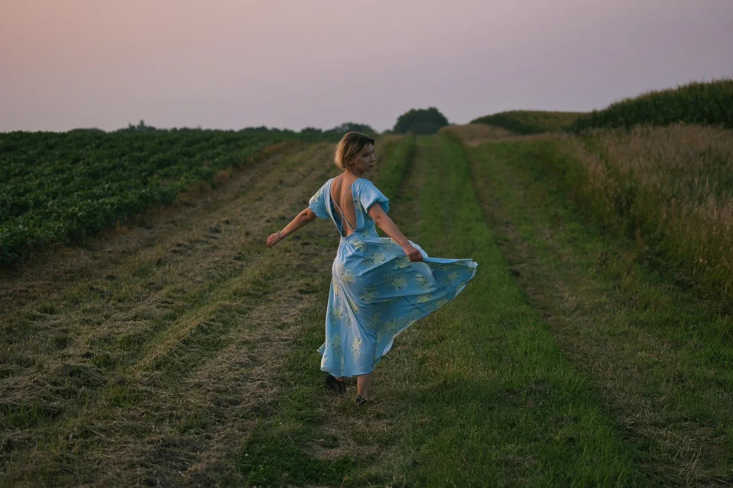 A woman in a blue dress with yellow floral patterns walking on a grassy path between fields at sunset.