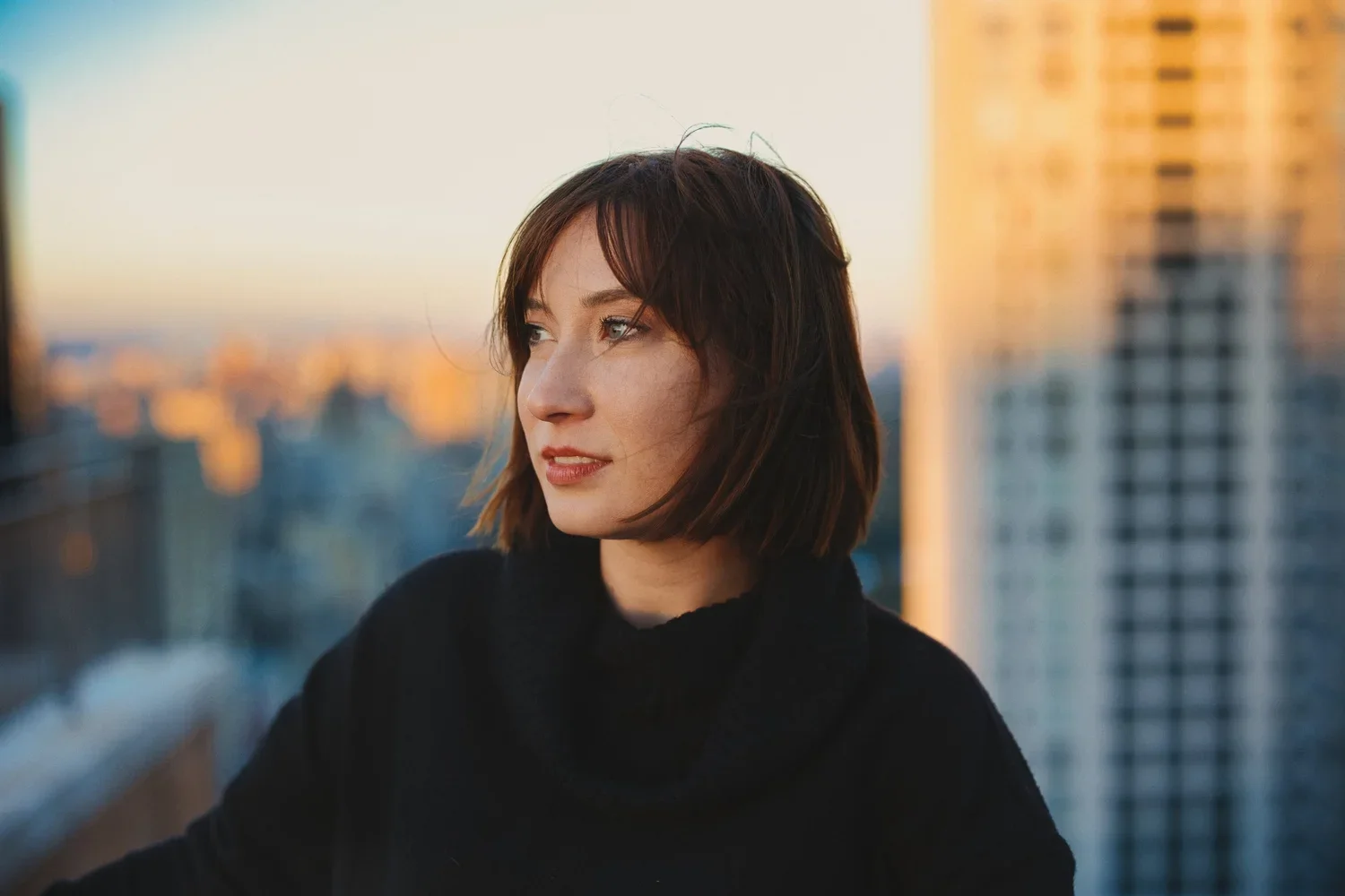A young woman with short, dark hair looking to the side on a city rooftop during sunset.