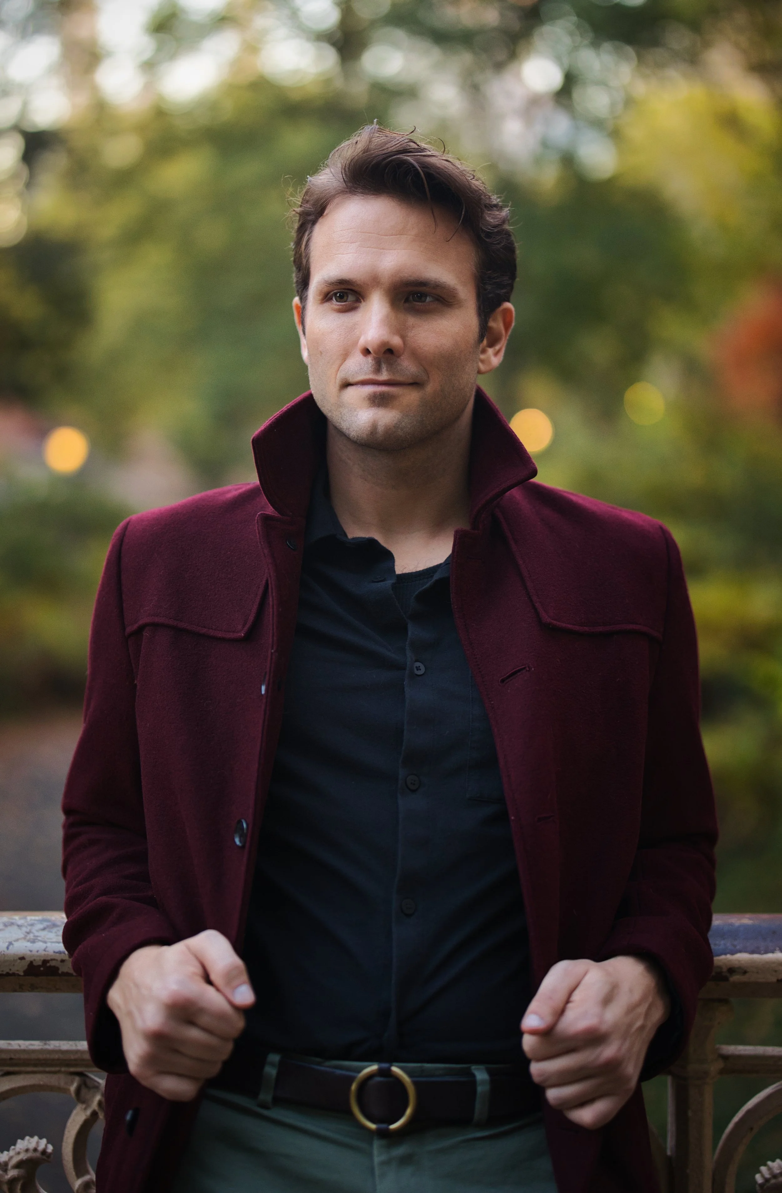 A man with dark hair wearing a maroon coat and black shirt is sitting on a park bench surrounded by autumn trees.