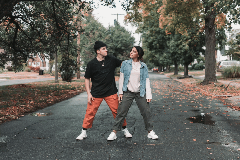 Two young people standing on an autumn leaf-covered street, facing each other with relaxed postures, surrounded by trees with fall foliage.