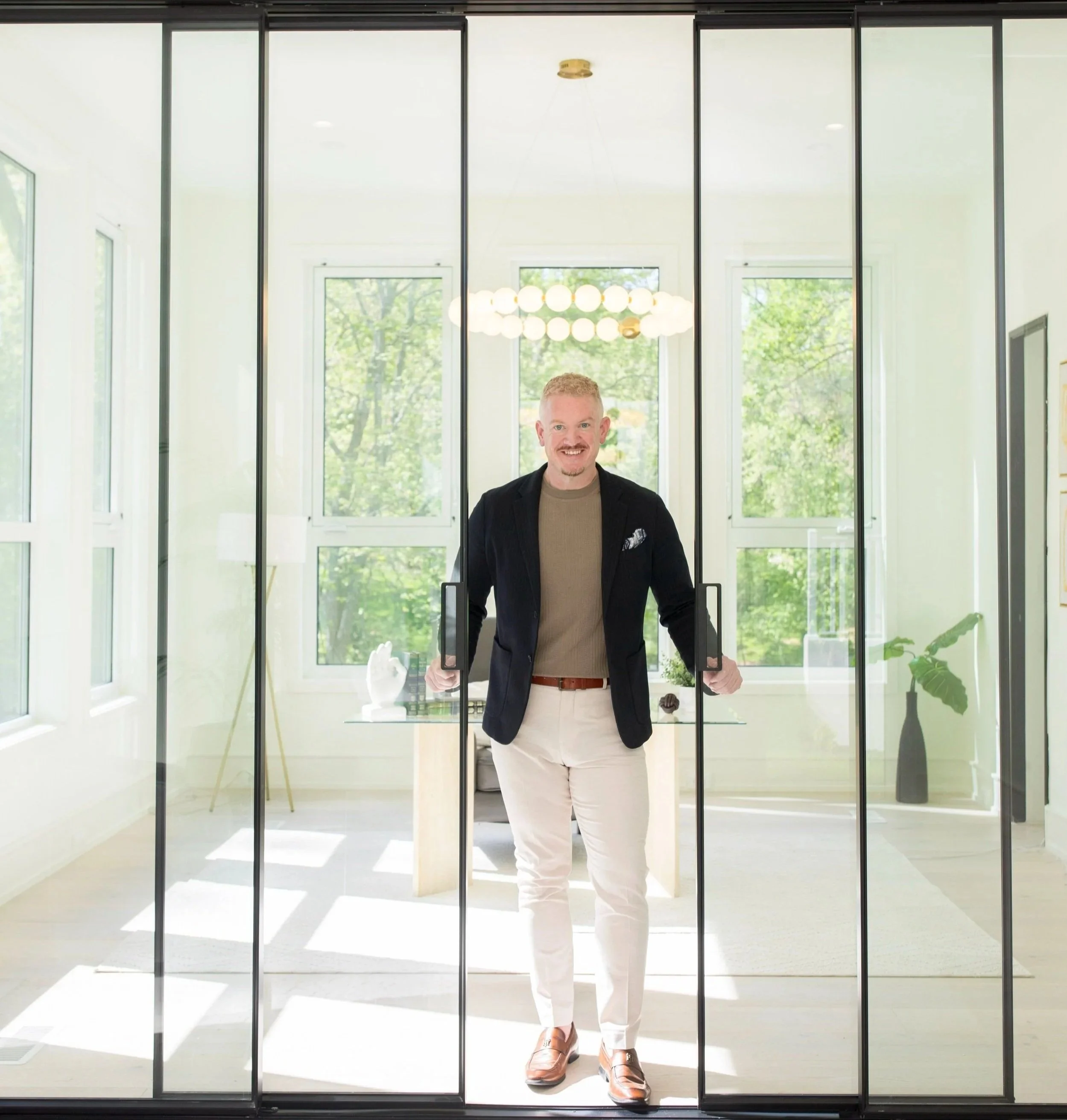 DC, MD, and VA Realtor Joshua Baumgardner in a blazer, brown sweater, and light-colored pants opening a glass door inside a bright room with large windows and green trees outside.