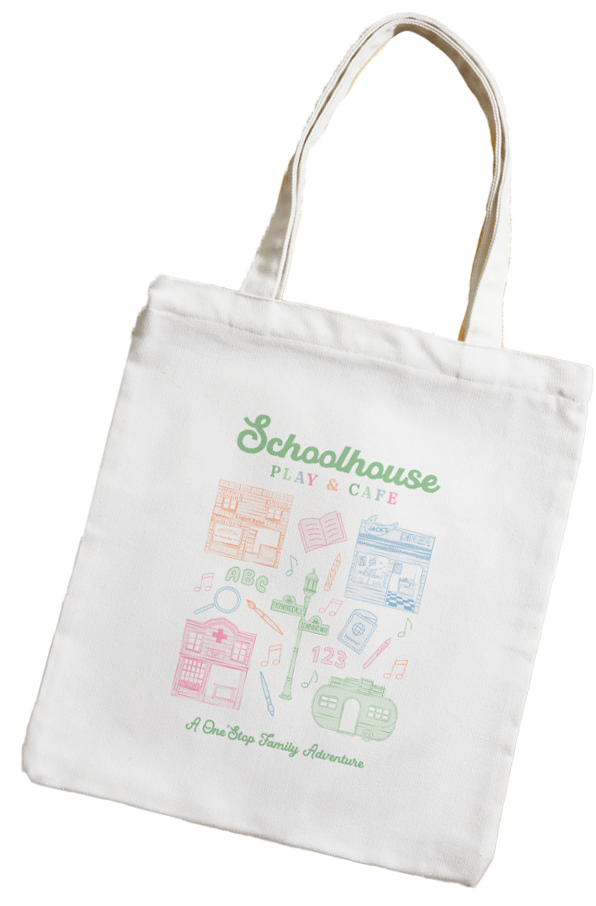 A white tote bag with colorful illustrations and text promoting a play and cafe for children, named "Schoolhouse Play & Cafe," featuring drawings of a schoolhouse, books, ABC letters, a clock, musical notes, and a family adventure slogan.