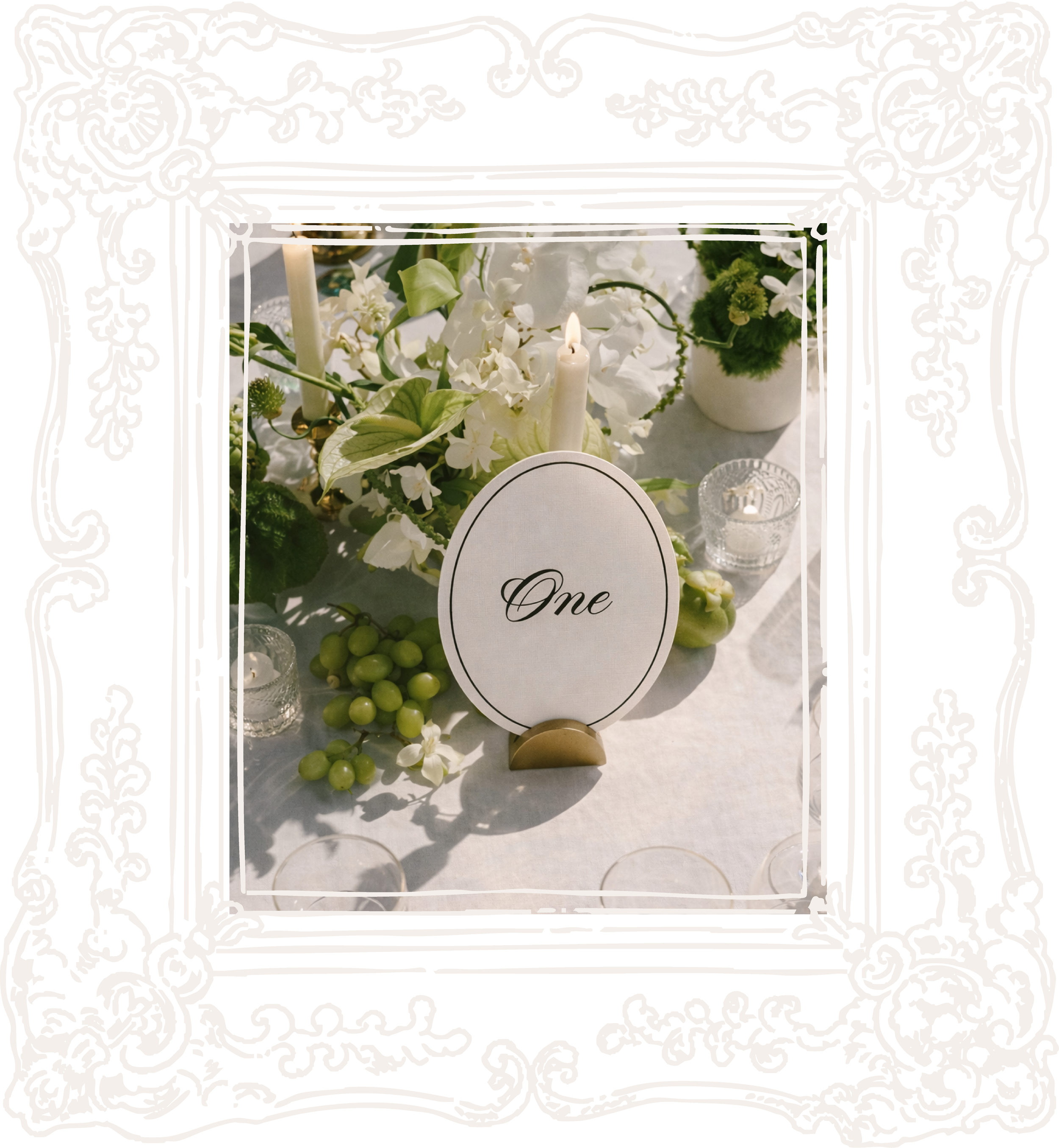 Table centerpiece with white flowers, green grapes, candles, and a sign that says 'One' for a wedding or celebration.