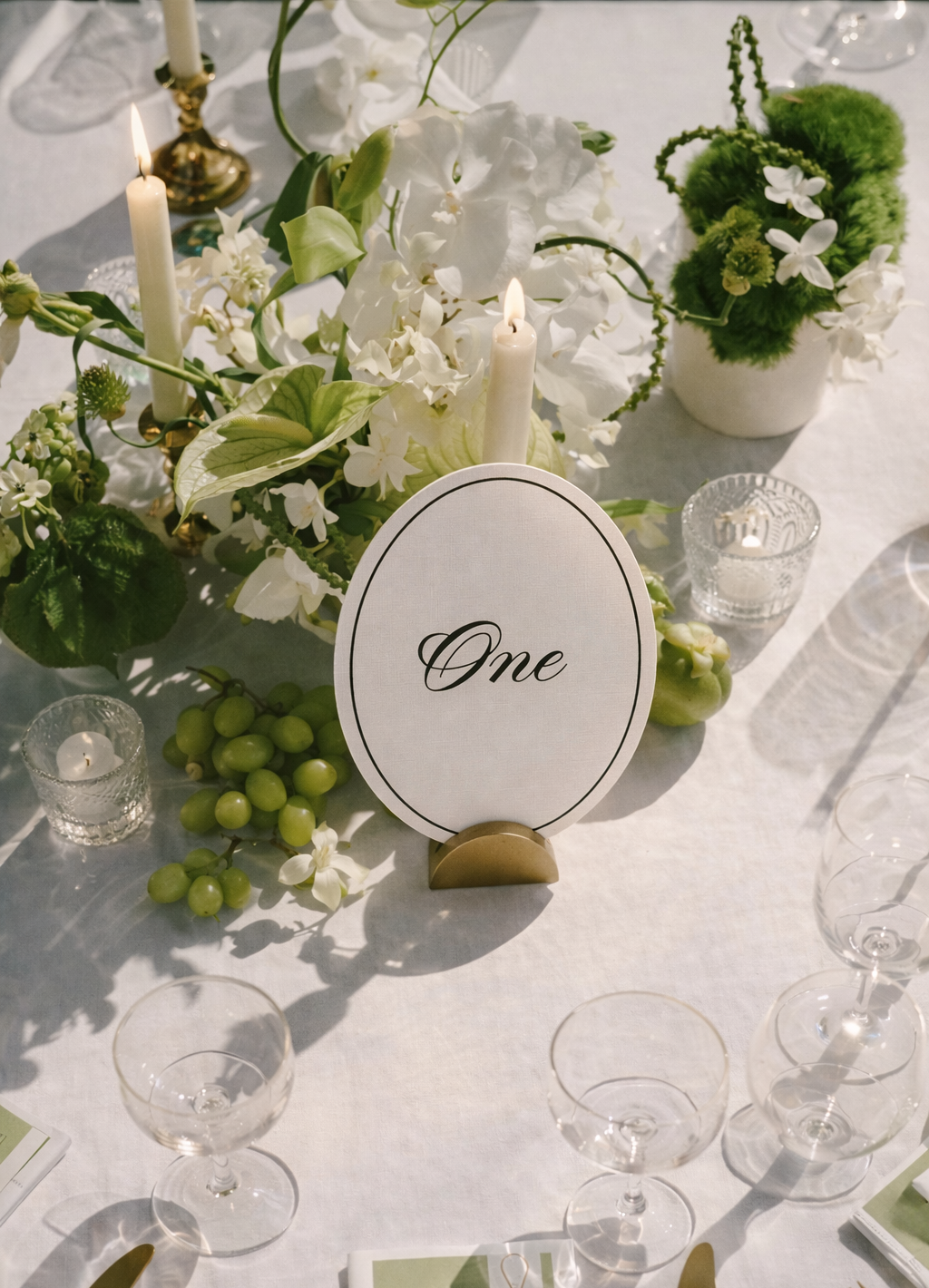 A close-up view of a table centerpiece for a celebration, featuring white flowers, glass candles, green grapes, a table sign that reads 'One', and decorative elements on a white tablecloth.