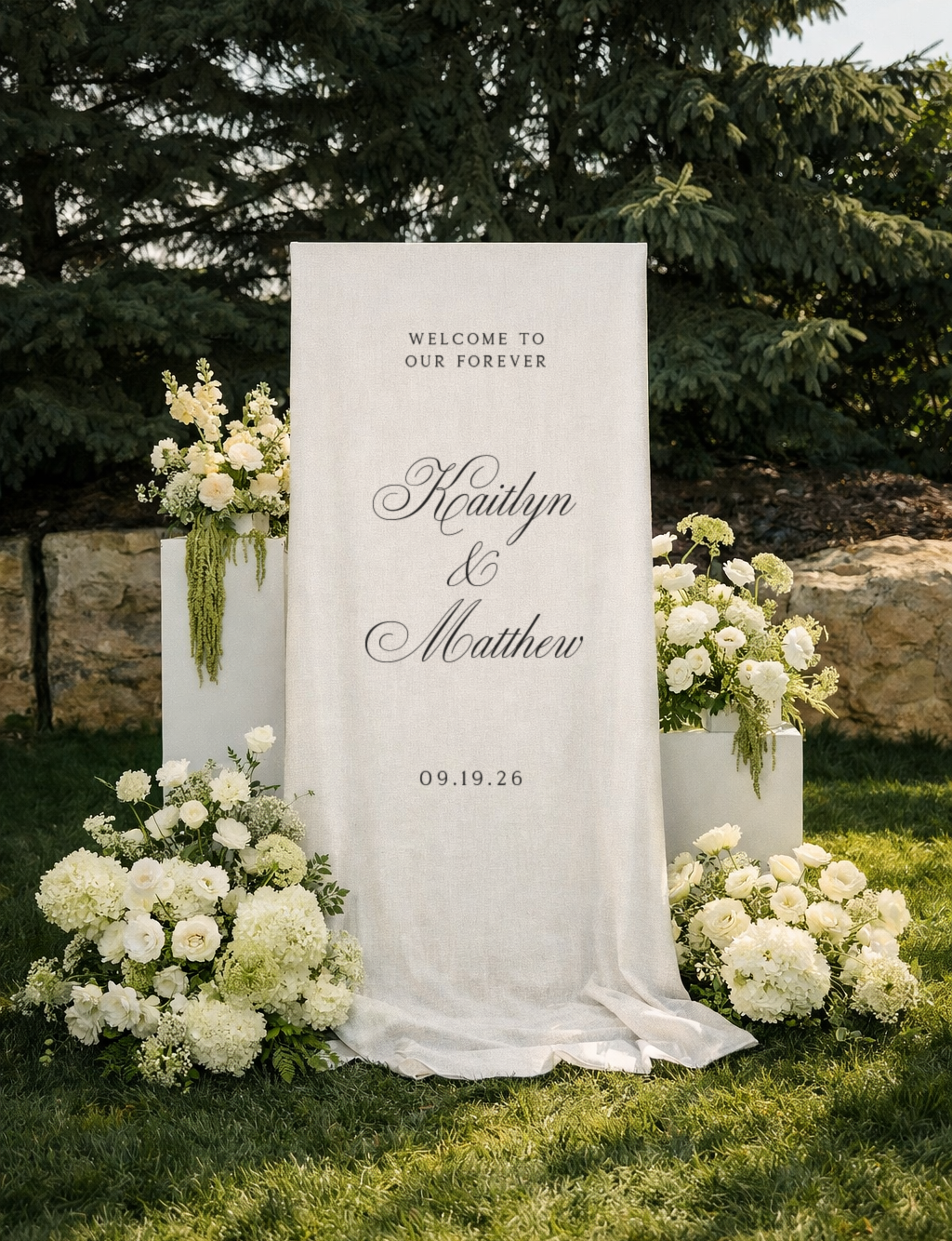 White wedding welcome sign with black cursive text, surrounded by white floral arrangements, outdoors on grass with trees and rocks in the background.