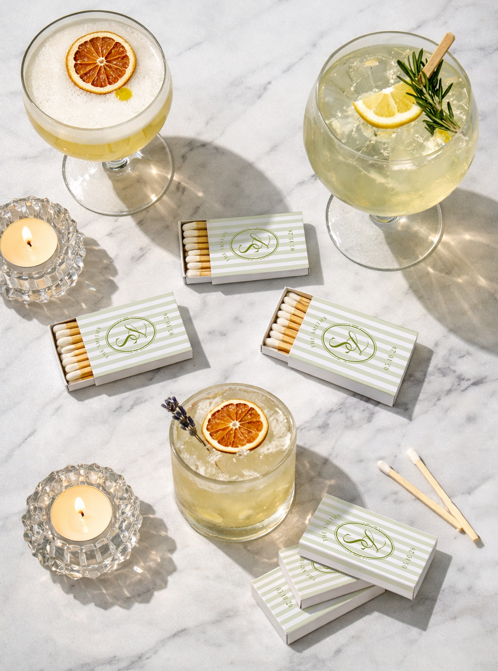 Two cocktails with lemon slices and herbs garnishes on a marble surface, surrounded by matchboxes and lit candles.