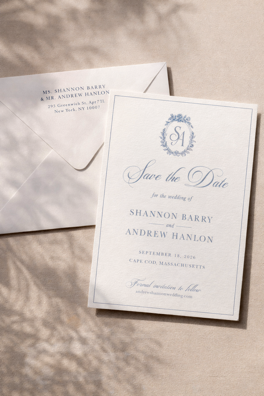 A wedding save the date card and an envelope on a beige surface. The card has elegant blue text and decorative elements, announcing the wedding of Shannon Barry and Andrew Hanlon scheduled for September 18, 2026, in Cape Cod, Massachusetts.