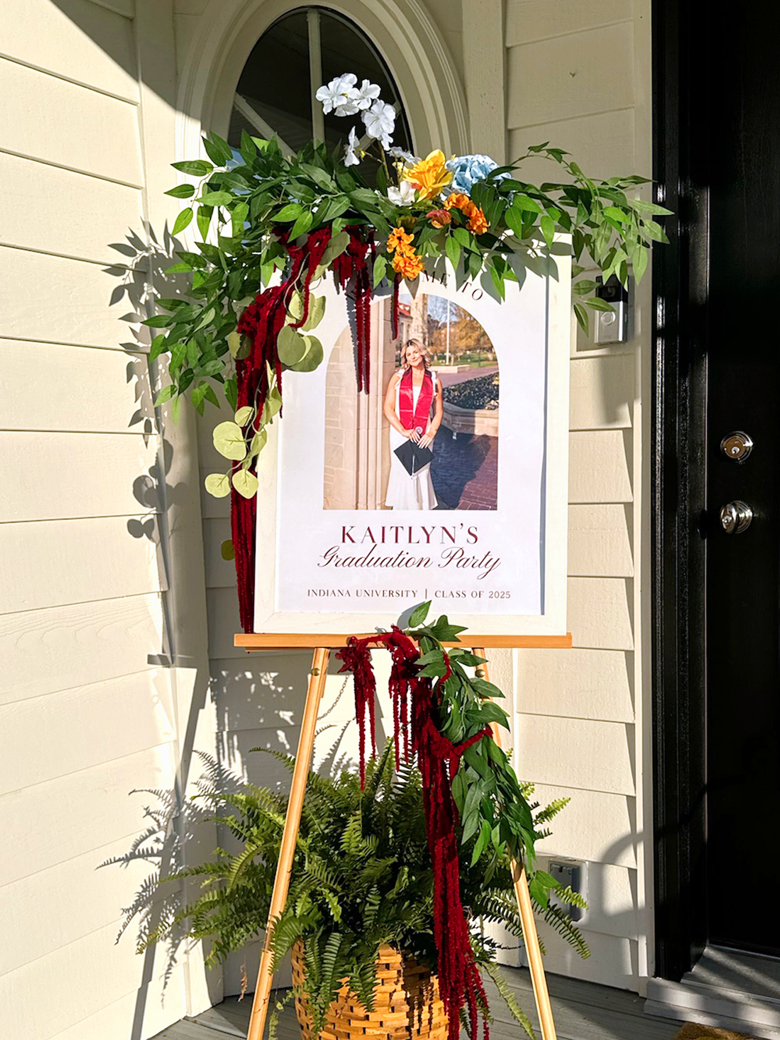 A welcome sign for Kaitlyn's graduation party features a photo of her in graduation attire. The sign is decorated with a floral arrangement of white, yellow, orange, and blue flowers and green leaves on an easel outside a house.