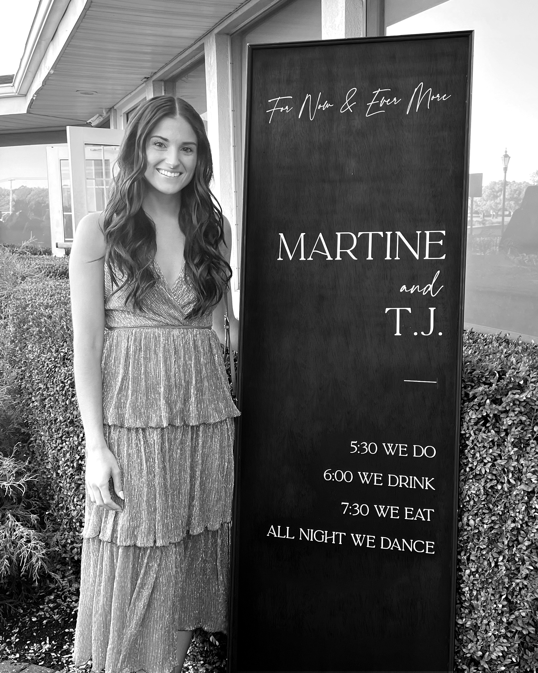 A woman in a long, shimmering dress standing next to a large blackboard sign outside a building.