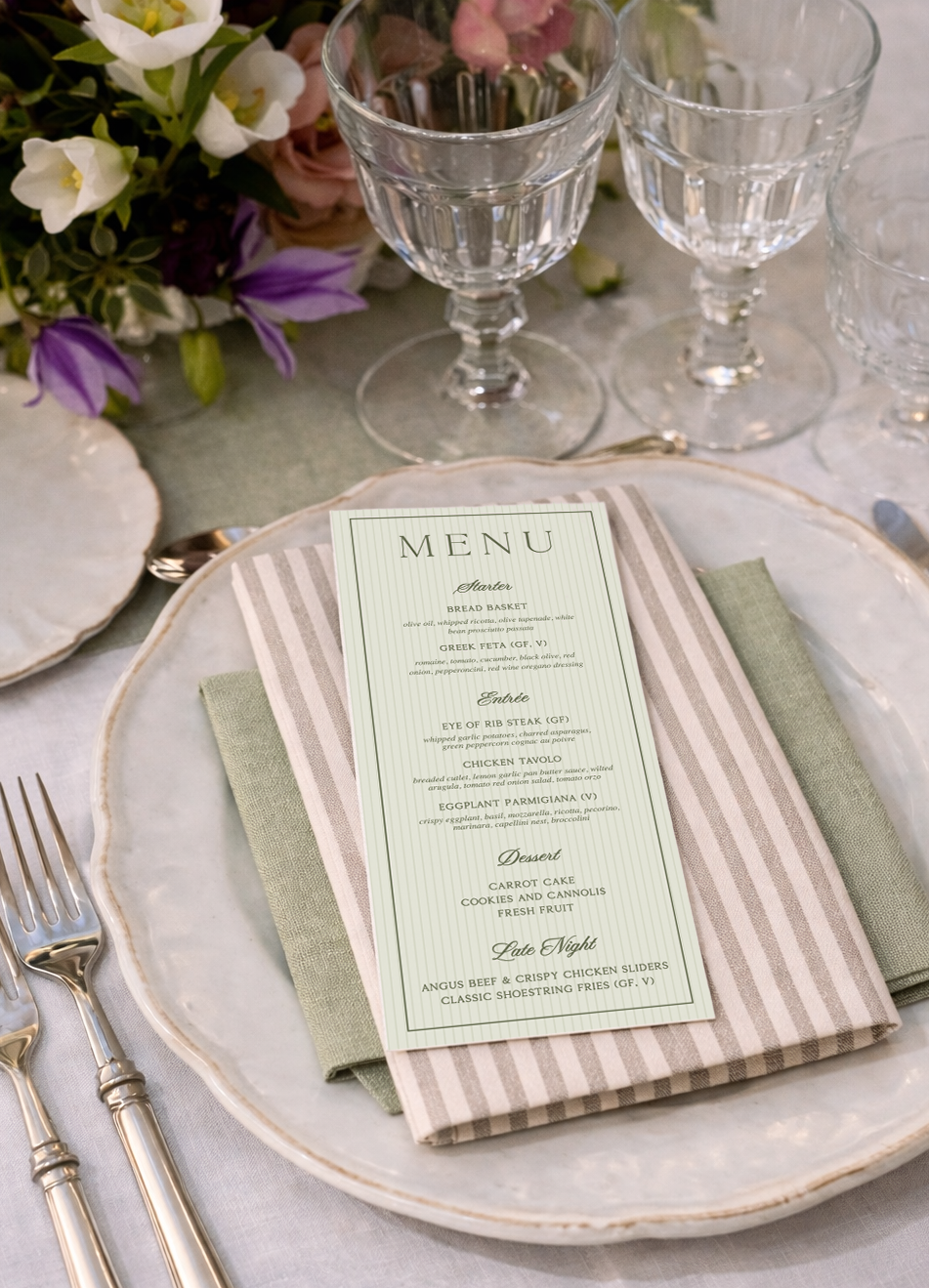 Elegant table setting with a menu on a layered napkin, surrounded by glassware and a floral centerpiece.