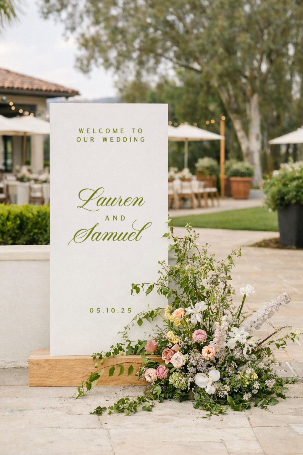 Wedding welcome sign with floral arrangement outdoors, with tables, umbrellas, and trees in the background.