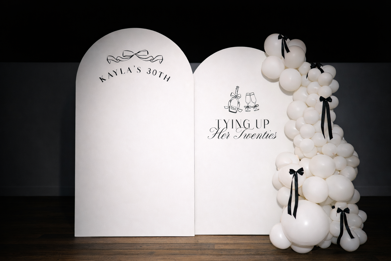 White backdrop with black text and graphics, decorated with white balloons and black ribbons. The text reads 'Kayla's 30th' and 'Tying Up Her Twenties' with champagne and bow illustrations.