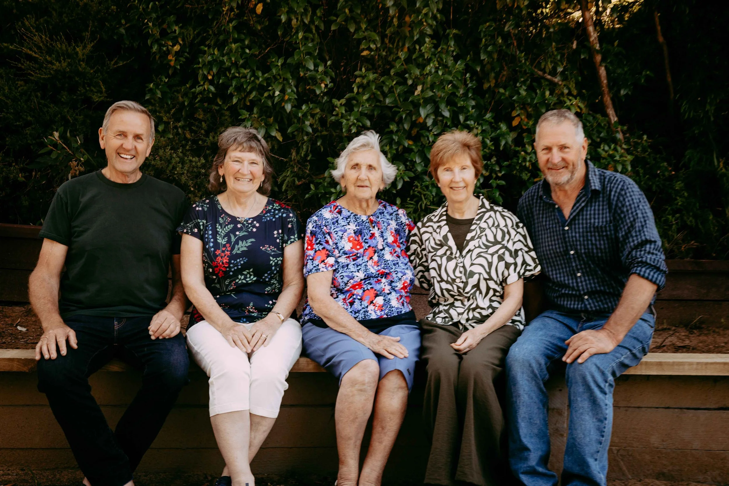Five older adults sitting on a wooden bench outdoors in front of green foliage, smiling at the camera.