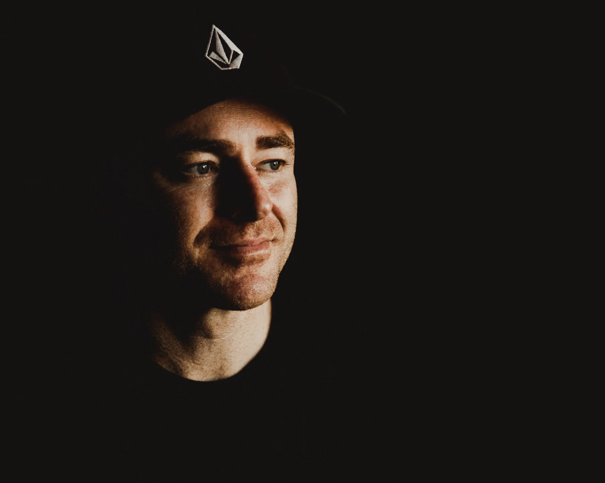 A close-up portrait of a young man wearing a black cap with a logo, illuminated by soft light, with a dark background.