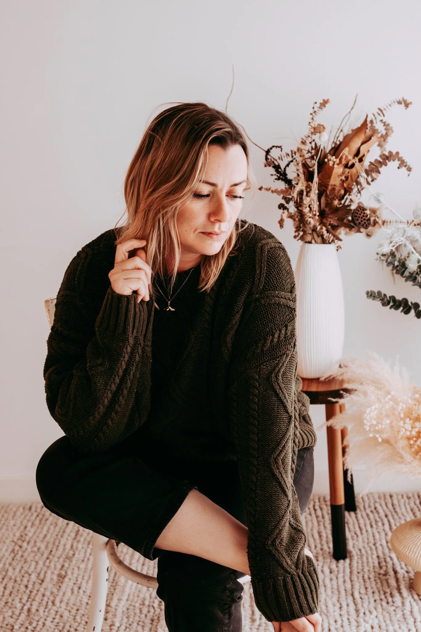A woman with shoulder-length blonde hair wearing a dark sweater and black pants, sitting on a chair in a room with neutral decor, next to a tall white vase with dried flowers, and a small wooden table with more dried flowers and plants.