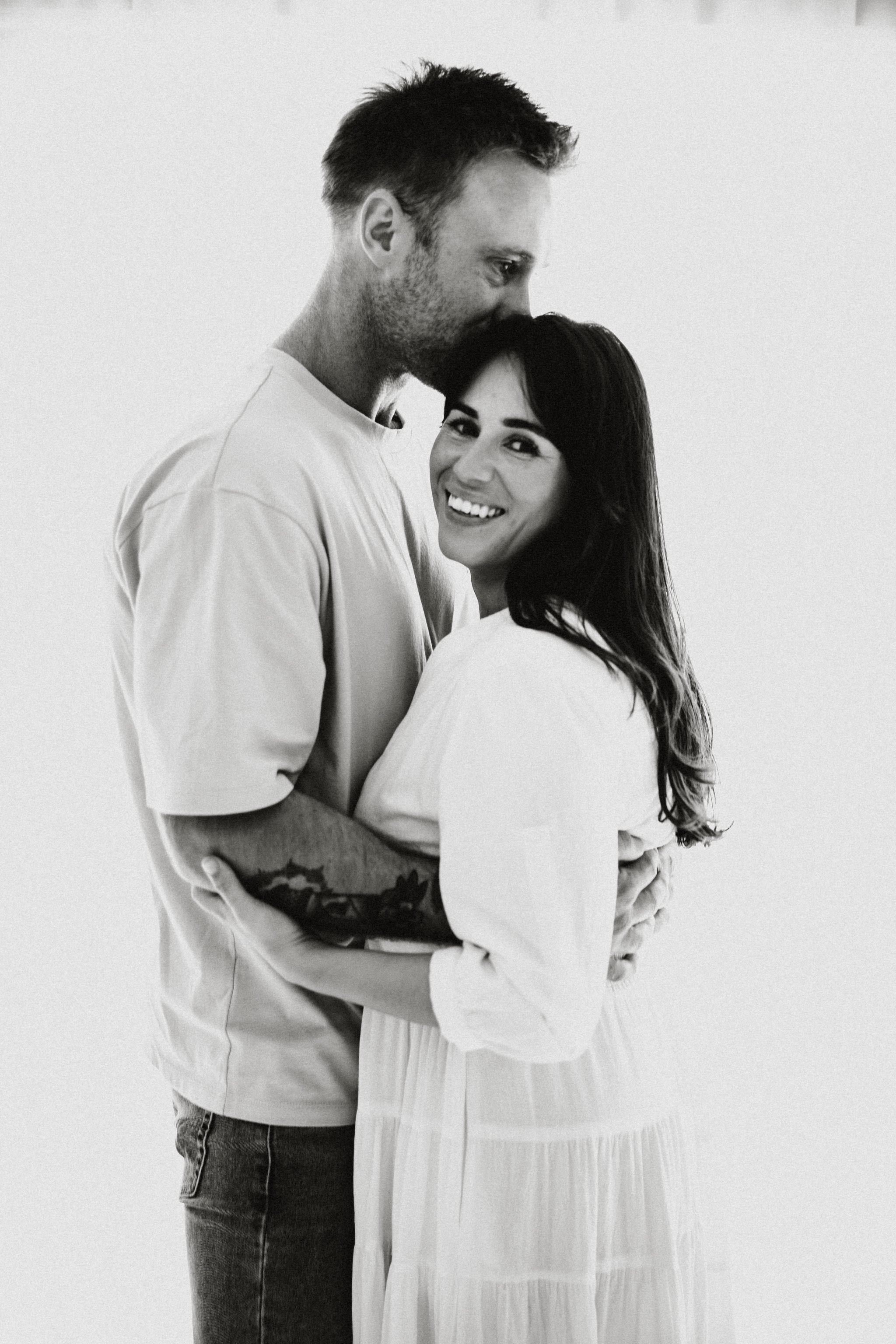 A happy couple embraces, with the man kissing the woman's forehead and the woman smiling at the camera, in black and white.