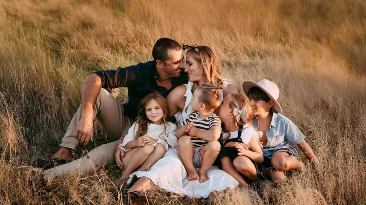 A family of six sitting in a field of tall grass, touching foreheads and smiling at each other during sunset.