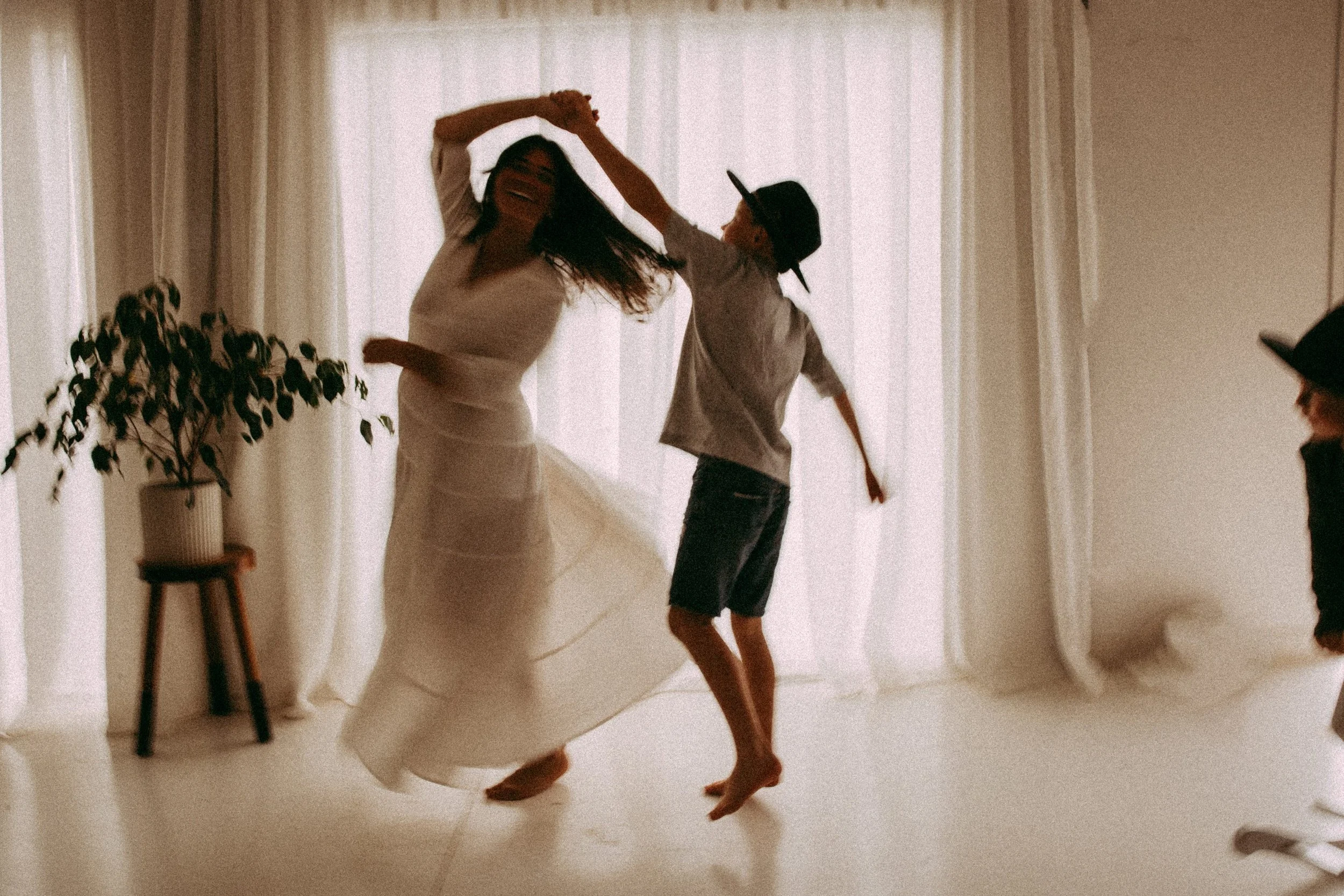 Woman dancing with two children, one boy, in a room with sheer curtains, a potted plant, and soft natural light.