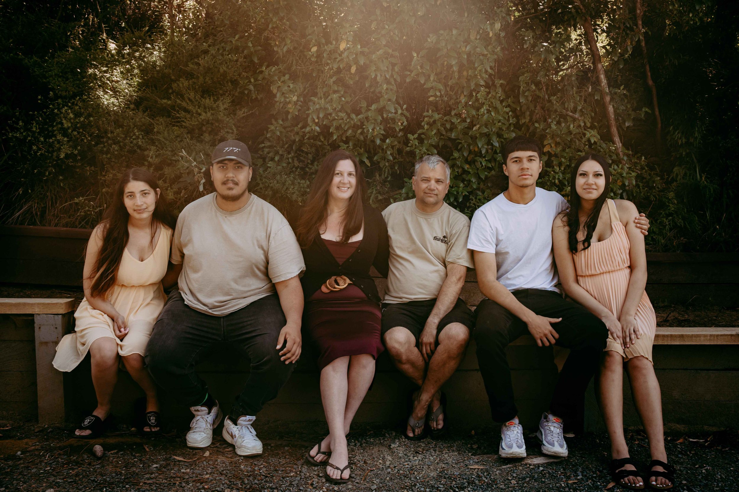 Group of six people sitting on a wooden bench outdoors with trees in the background, some smiling and others looking serious, during the daytime.
