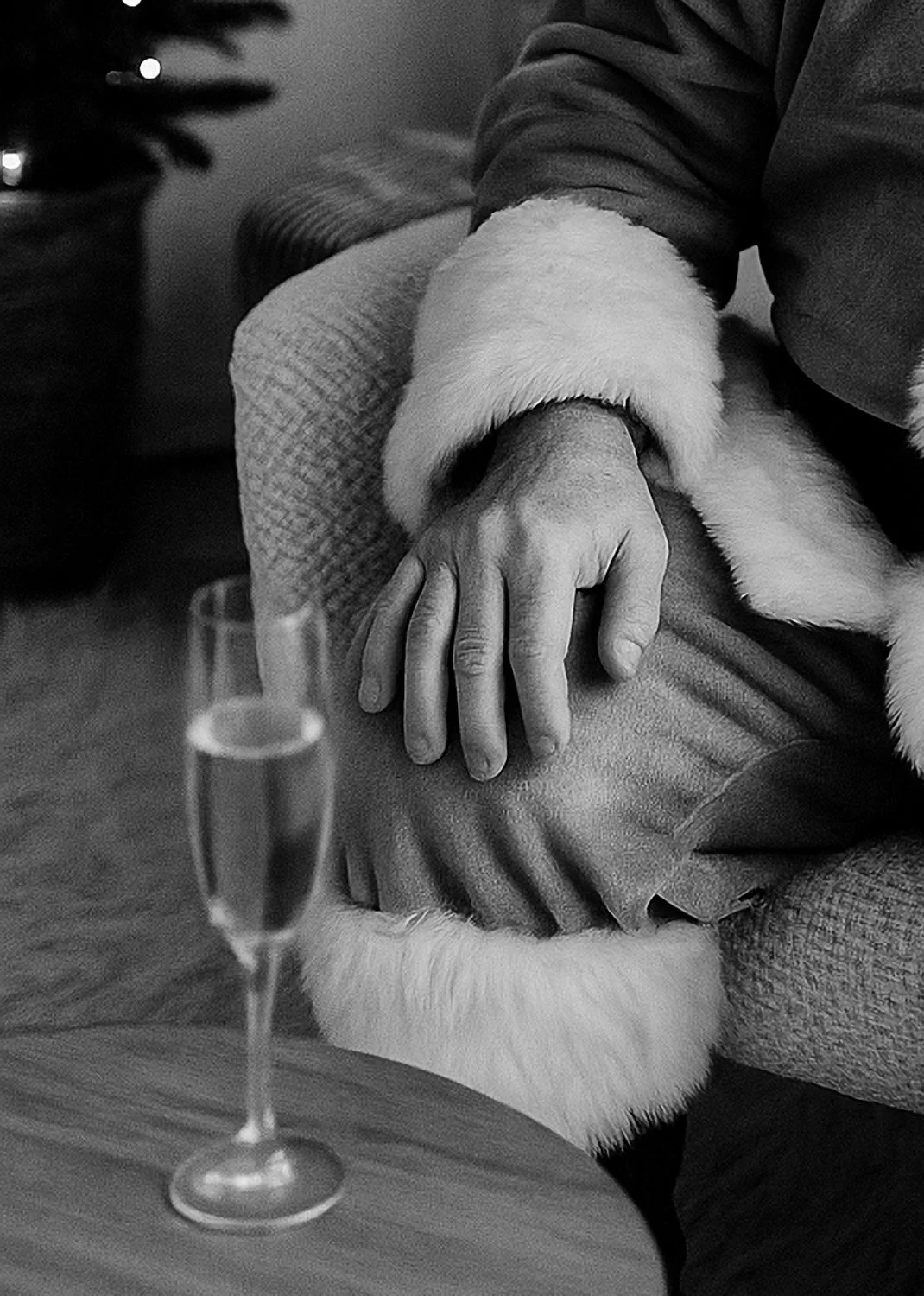A person's hand resting on their knee, which is covered with a fluffy white fur cuff, in a cozy setting. There is a glass of champagne on a table in the foreground, and a decorated Christmas tree in the background.