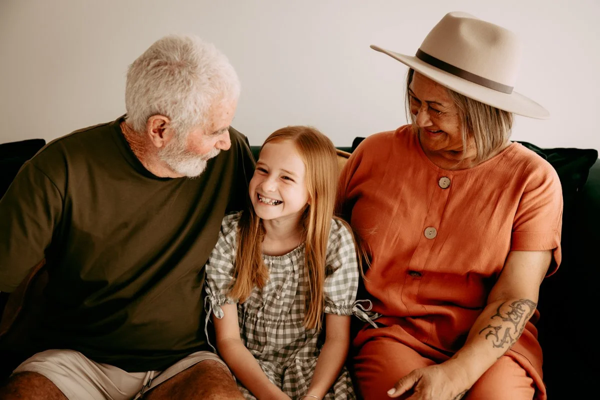 An elderly man with white hair and a beard, wearing a dark t-shirt, sitting next to a young girl with long red hair, wearing a checked dress, and an elderly woman with a wide-brimmed hat and tattooed arm, wearing a rust-colored dress; all three are smiling and looking at each other, sitting on a black sofa against a plain background.