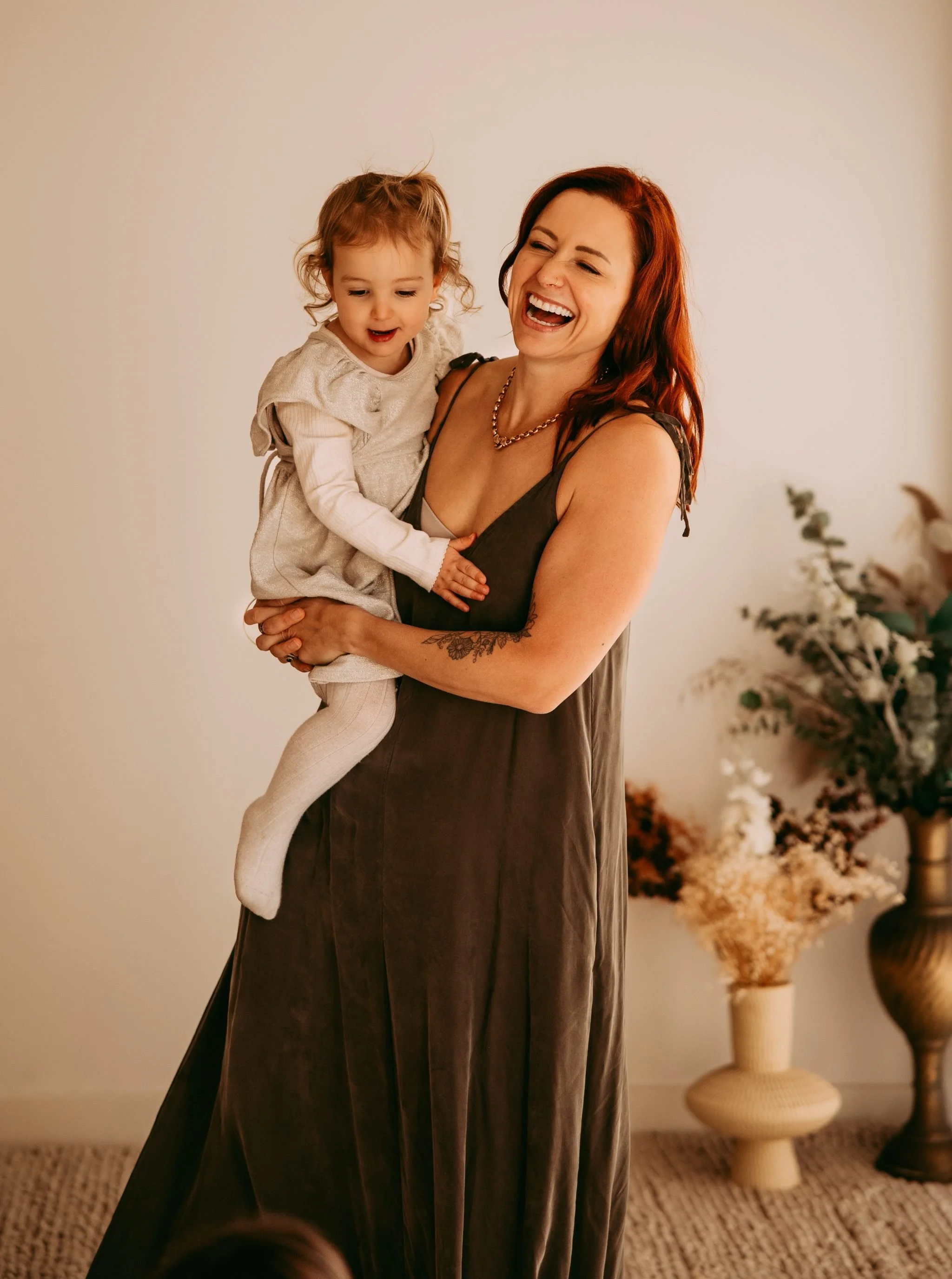 A woman with red hair holding a young girl with curly hair, both laughing, indoors with neutral wall and vases of dried flowers in the background.