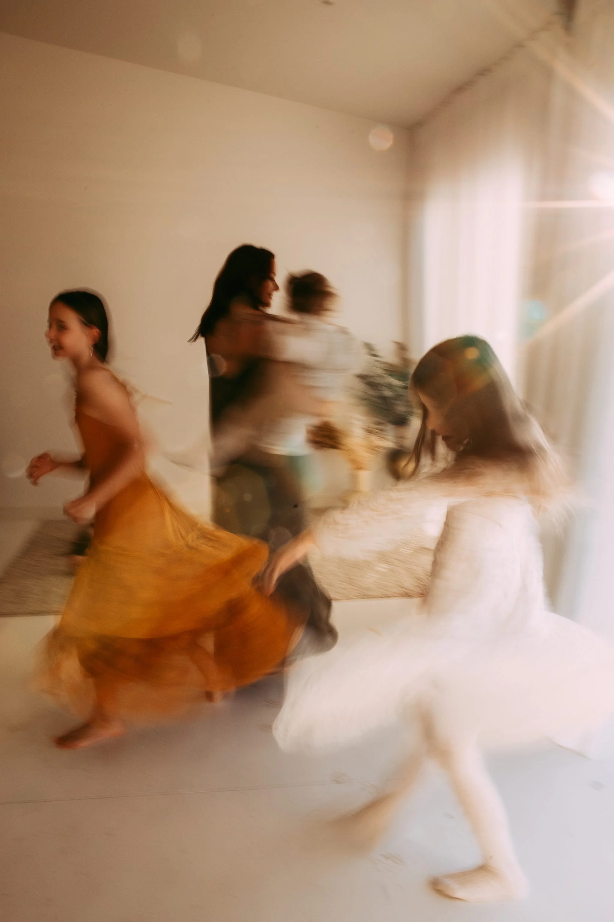 Blurry photo of two women and three children dancing indoors, with sunlight coming through a window, and a dog in the foreground.