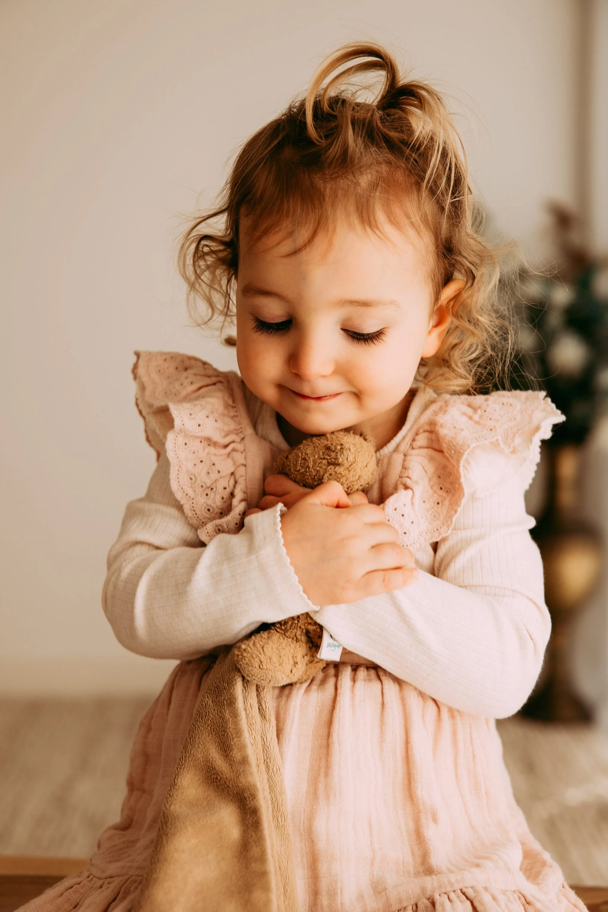 A young girl with curly blonde hair hugging a stuffed animal close to her chest, with her eyes closed and a gentle smile.