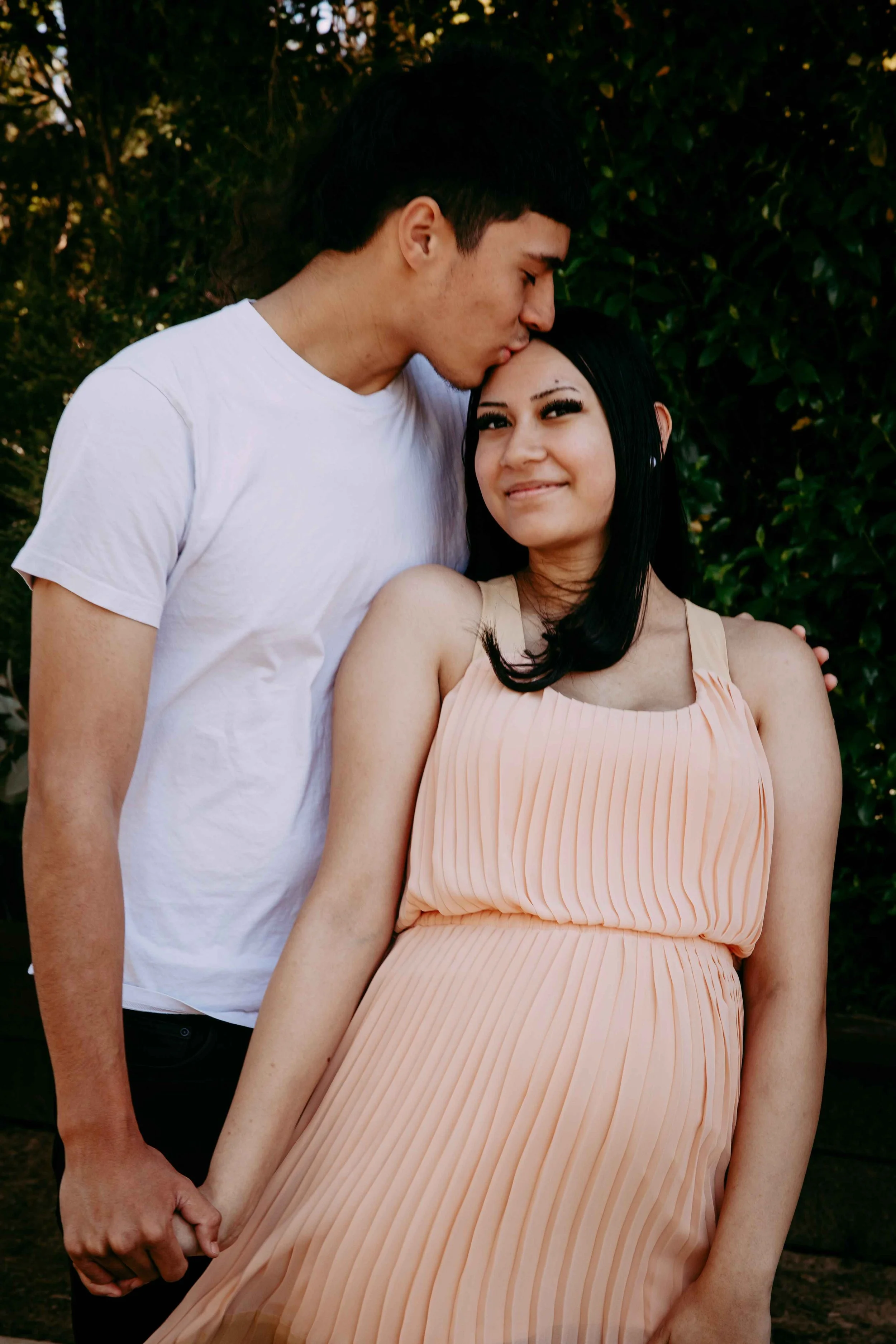A young couple holding hands outdoors, with the man kissing the woman's forehead and the woman smiling, in front of green bushes.