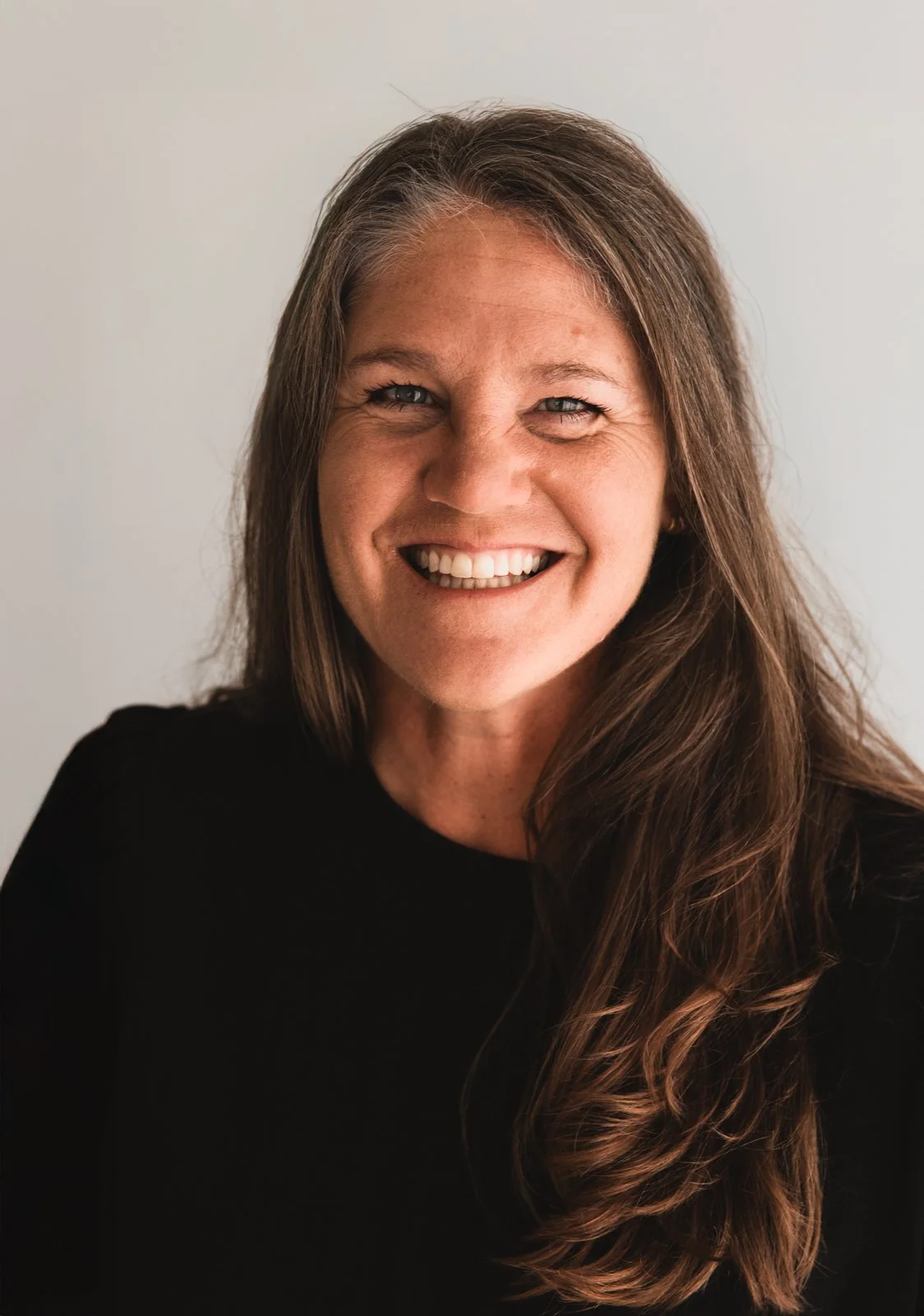 Smiling woman with long brown hair wearing a black top, against a neutral background.