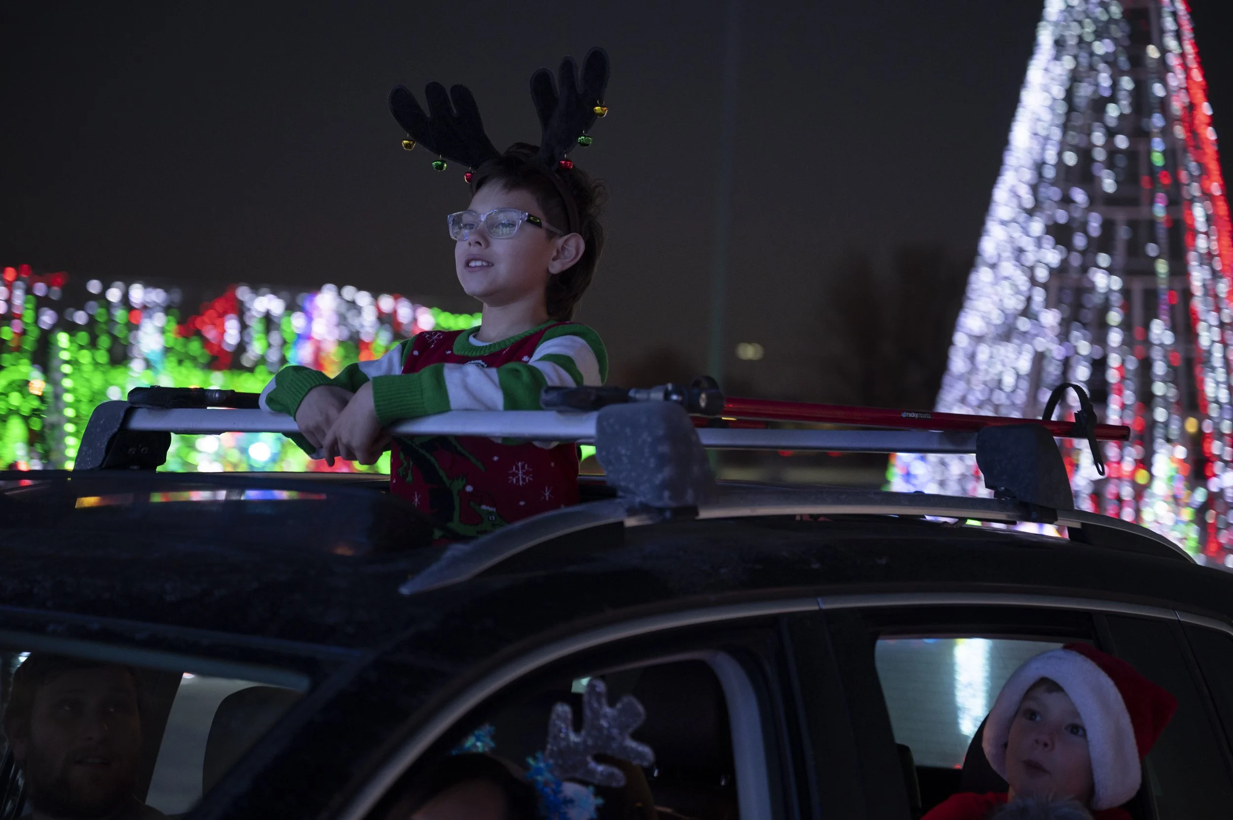 A boy wearing reindeer antlers with Christmas lights, glasses, and a festive sweater rides in a car decorated for Christmas. Two children are in the car, one wearing a Santa hat. The background features brightly lit Christmas trees, with a large illuminated Christmas tree on the right side of the image.