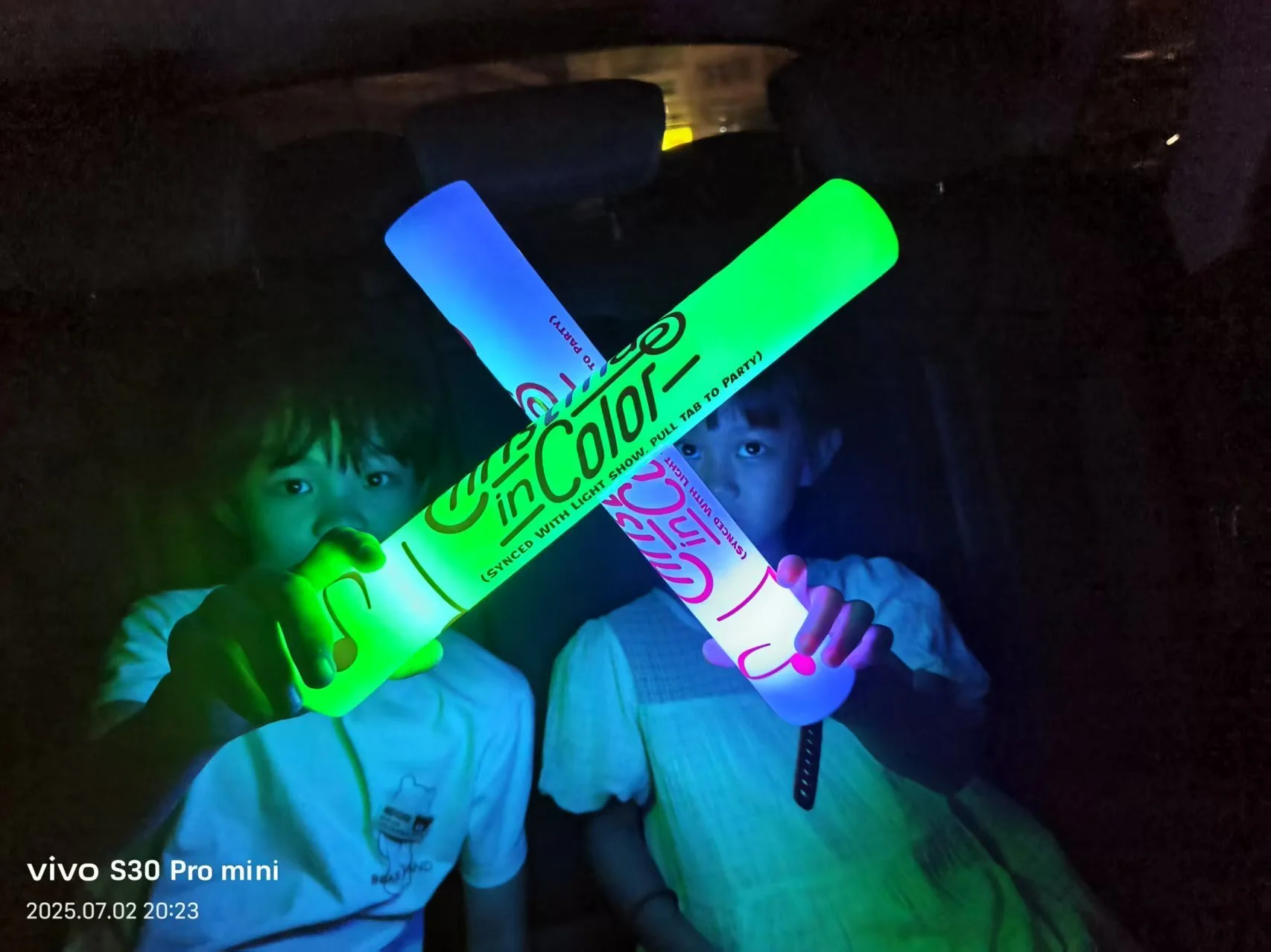 Two young boys holding glowing light sticks inside a dark room.