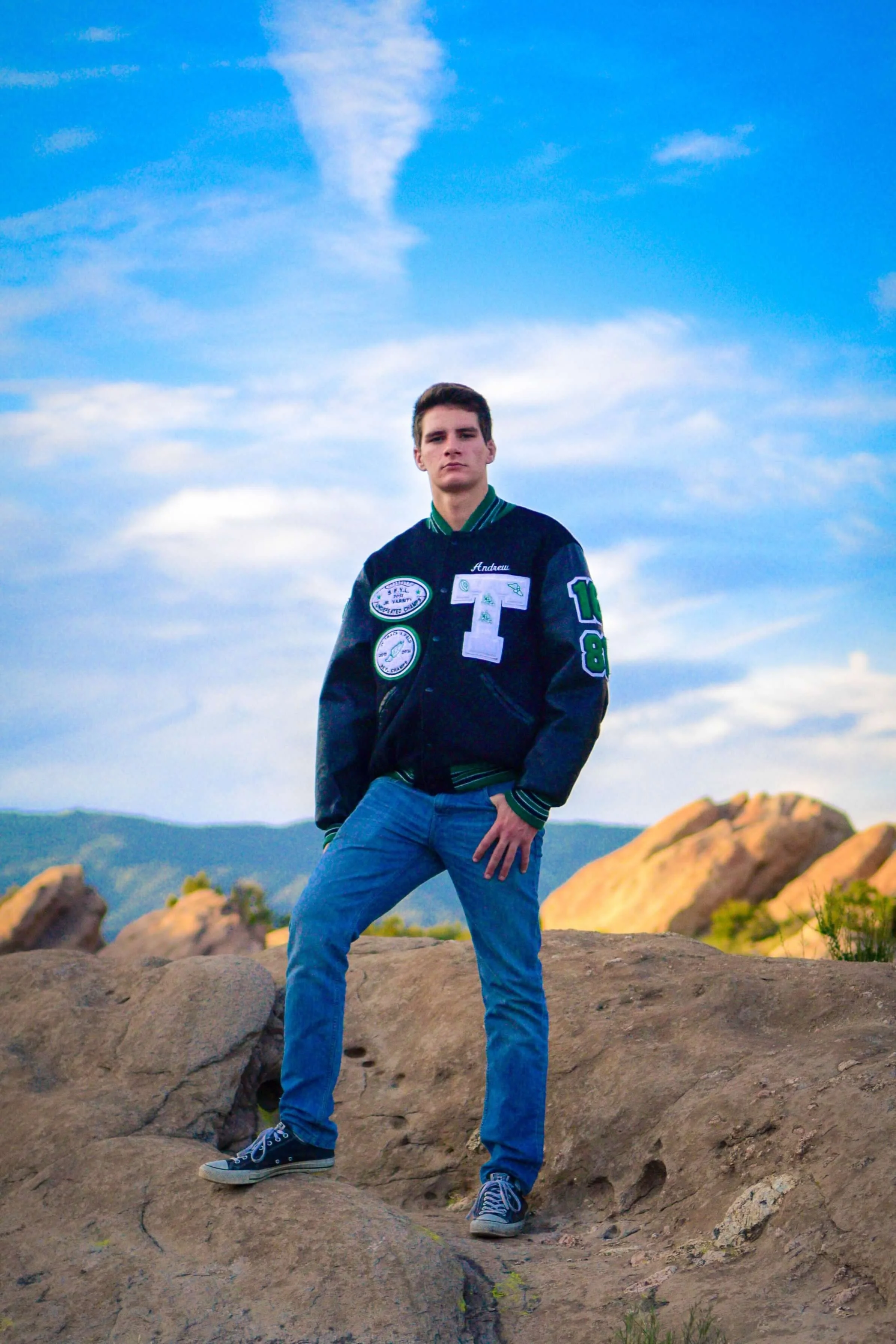 High school senior boy wearing varsity football jacket, standing with feet apart, looking strong and seriously toward camera, with Vasquez Rocks in background.