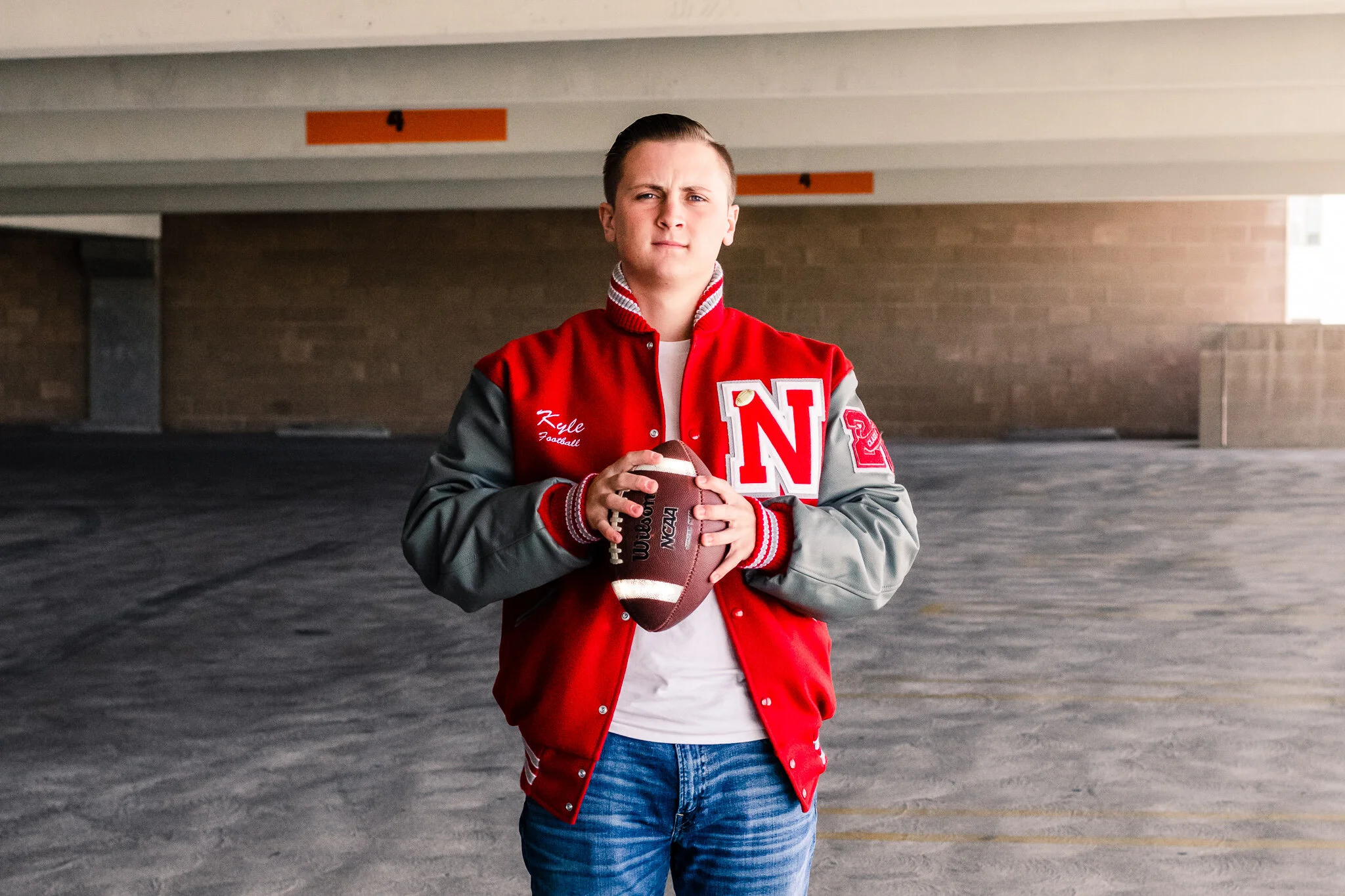 Senior guy wearing red letterman's jacket holding a football, looking straight at camera.