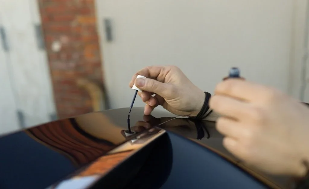 Close-up of a hand using a small brush to apply touch-up paint into a chip on a car, showing precise, careful application.