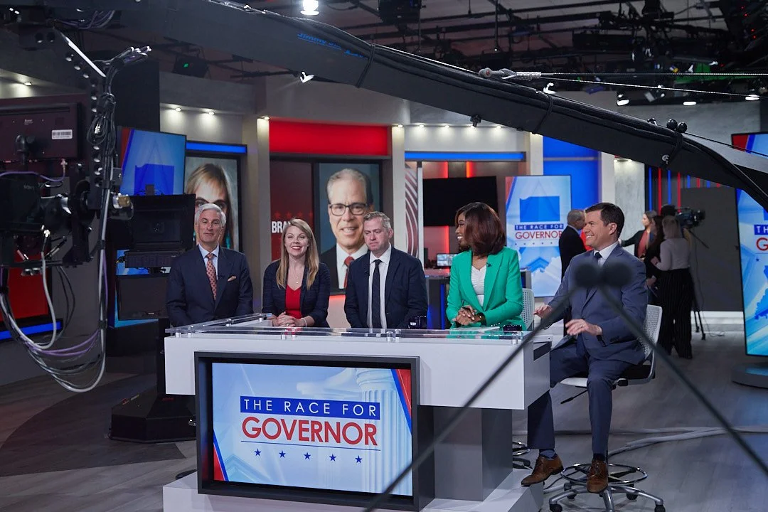 Television news studio with five diverse candidates seated at a table, discussing for a gubernatorial debate, with campaign posters and screens in the background.
