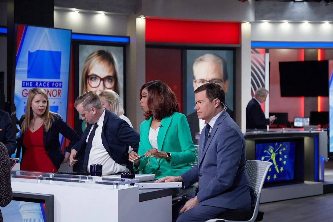 Television news studio with hosts and guests discussing, in front of political debate screens.