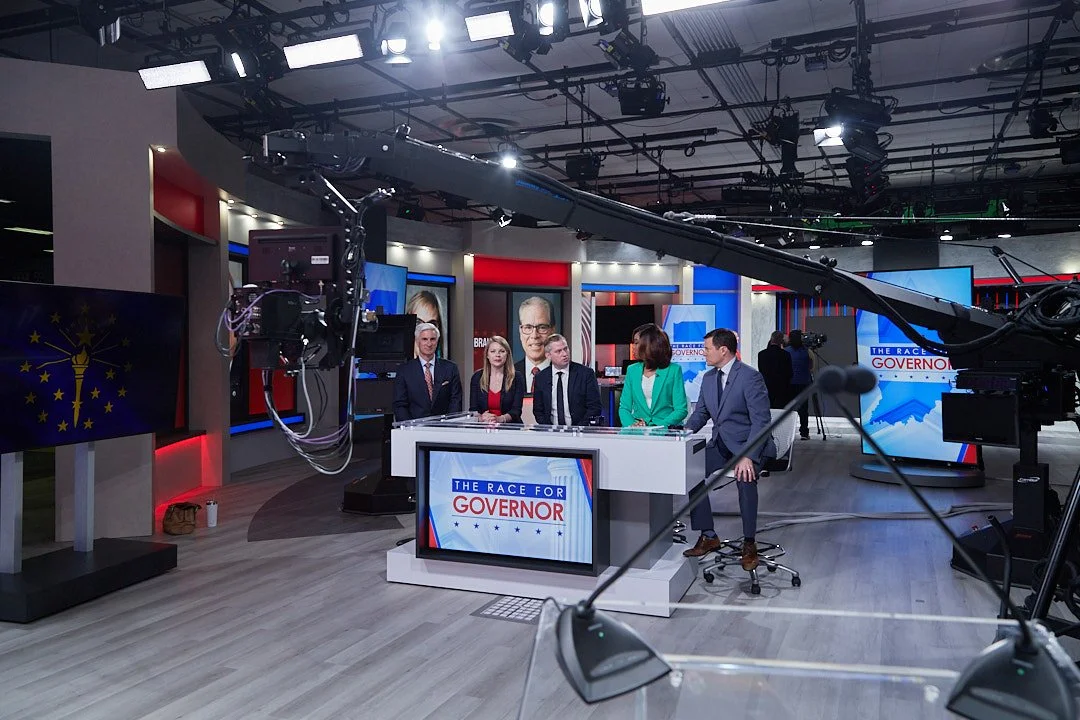 Television news studio with five news anchors sitting at a desk, studio equipment, cameras, and large screens displaying graphics related to the race for governor.