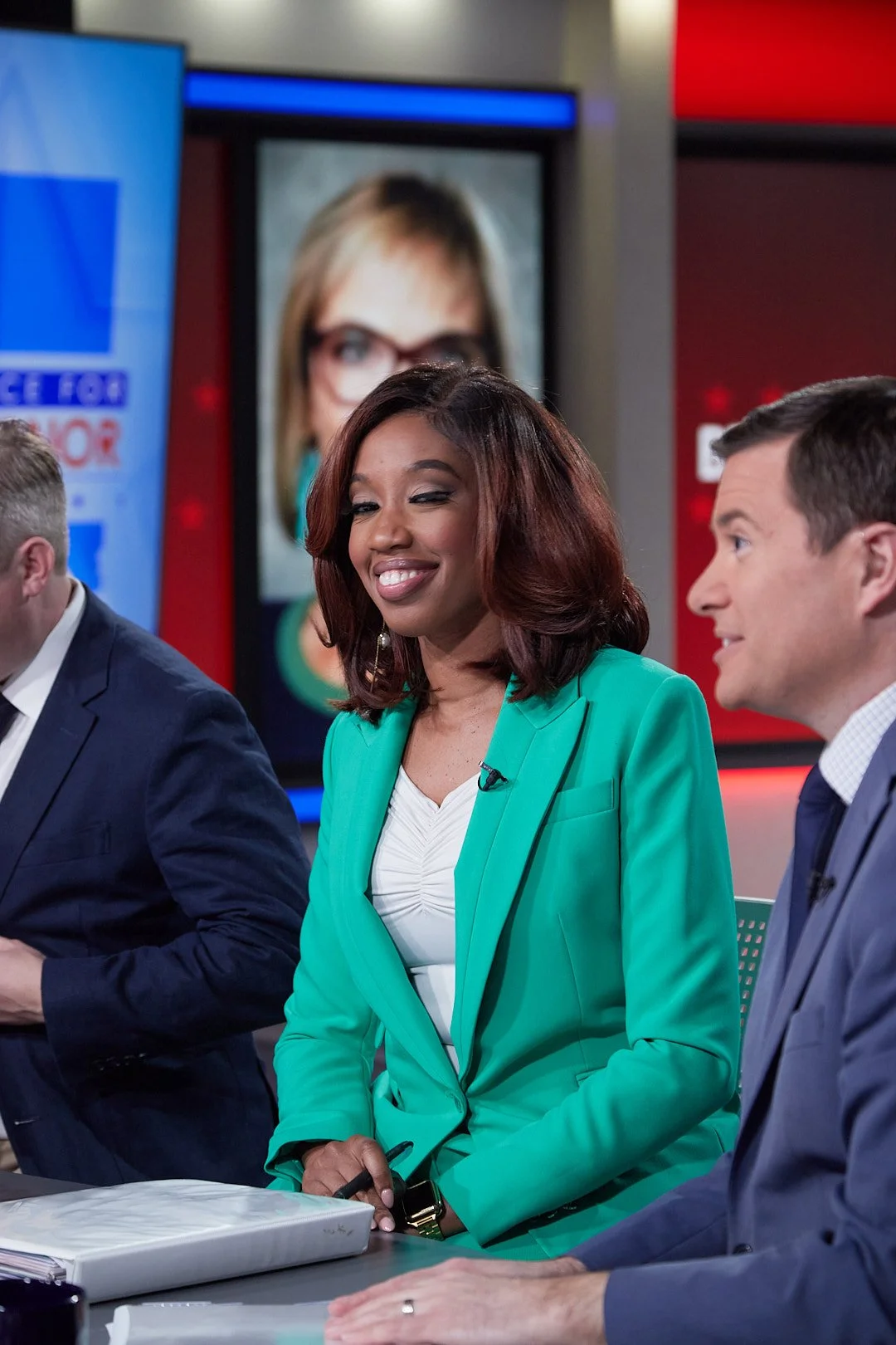 A woman in a teal blazer sitting at a news desk, smiling with a television studio background.