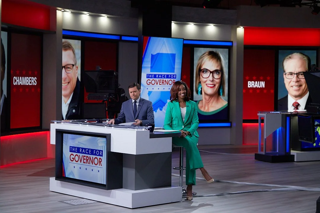 Television news studio with two anchors, a man in a gray suit and a woman in a green suit, sitting at a desk with a screen that reads "The Race for Governor." Large screens in the background display images of two men and their names, "Chambers" and "