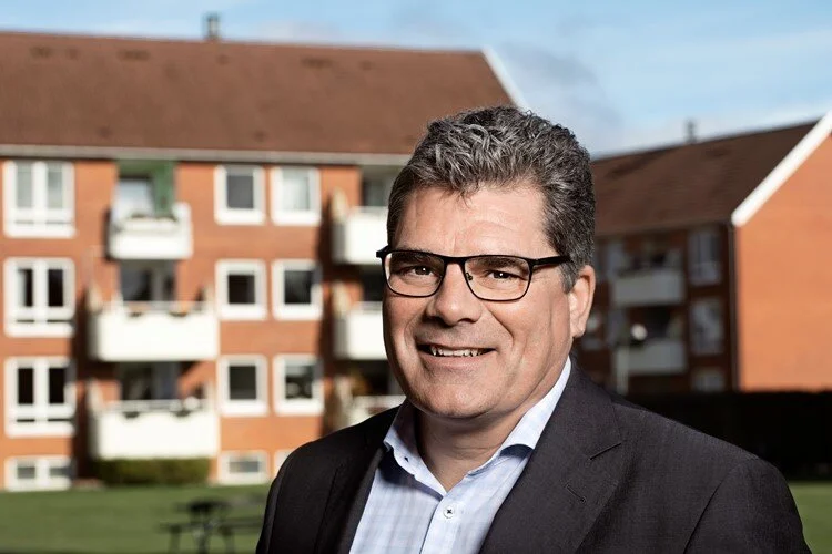 Mand iført briller og et jakkesæt, der står udendørs med murede lejlighedsbygninger i baggrunden