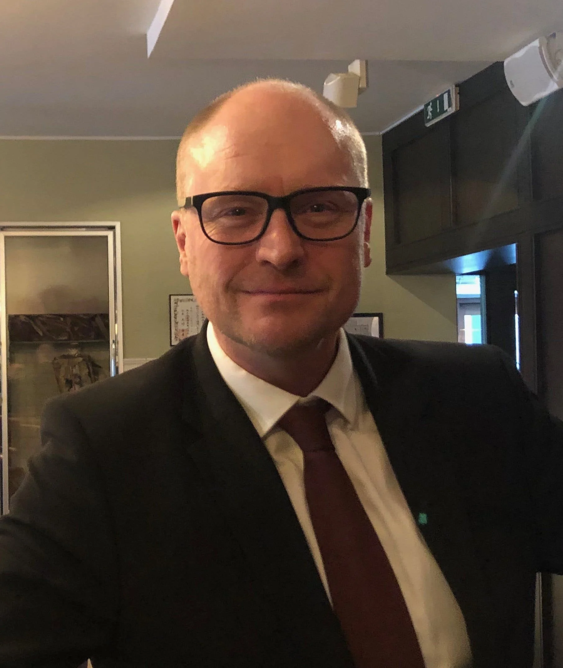 En mand iført briller og et jakkesæt med slips, stående indendørs med et neutralt udtryk.