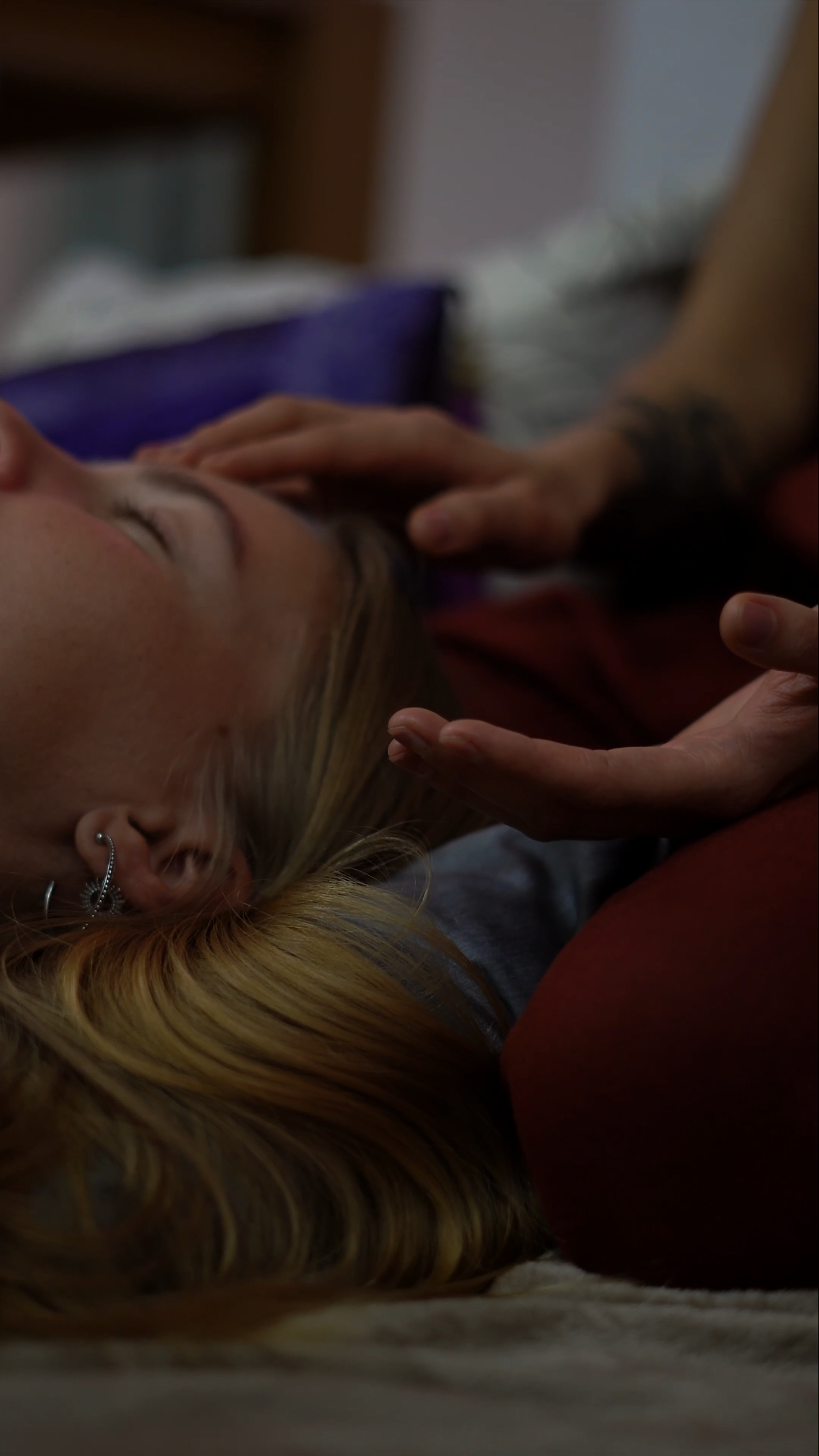 A light-skinned woman with blonde hair lying down with eyes closed, as a person with a tattooed arm gently touches her head.