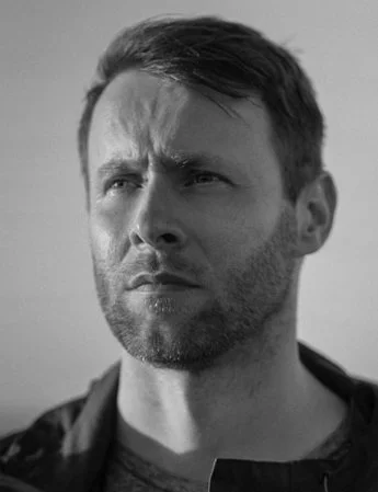 Black and white photo of a man with short hair and a beard, looking to the side with a serious expression.