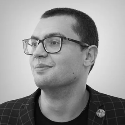 Black and white photo of a man with glasses, short dark hair, wearing a checkered blazer and a pin on the lapel, looking slightly to the side.