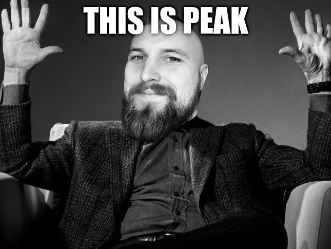 A man with a beard and a mischievous smile sitting with his arms raised, in a black-and-white photo, with the text "THIS IS PEAK" above his head.