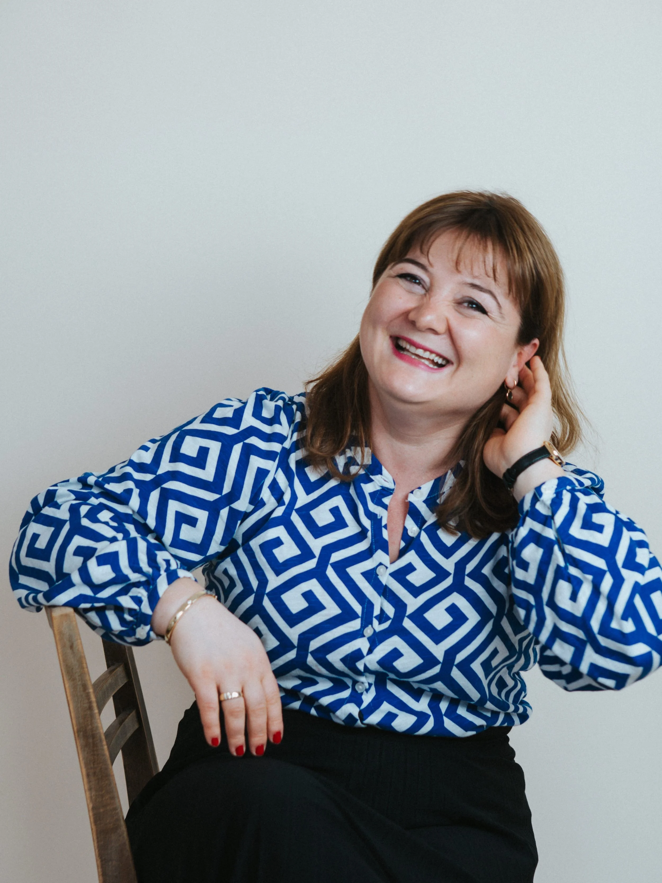 Frau im blauen und weißen Musterhemd, lächelnd, sitzend an einem Stuhl, Blick gerecht, mit braunen Haaren, aufhellendem Hintergrund.