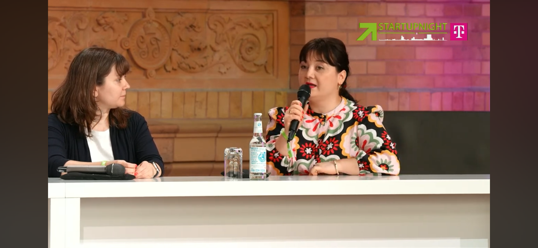 Zwei Frauen sitzen an einem Tisch bei einer Diskussionsrunde. Die Frau auf der rechten spricht in ein Mikrofon, während die Frau auf der linken zuhört. Es gibt Wasserflaschen und Gläser auf dem Tisch. Im Hintergrund sind gemauerte Wände und eine hölzerne Dekoration. Das Bild enthält ein Logo für 'STARTUPNIGHT' und eine Telekom-Partnerschaft.